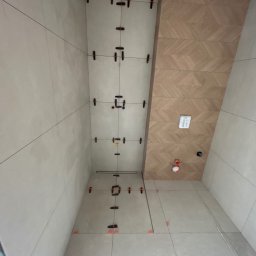 Firma ogolno budowlana Miroslaw Gros - Łazienka w trakcie wykańczania: jasne płytki na ścianach i podłodze, system poziomowania płytek, dekoracyjna ściana z drewnopodobnych płytek.