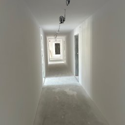 Firma ogolno budowlana Miroslaw Gros - Długi korytarz po gładziowaniu w Słopnicach. Widoczne gładkie, białe ściany, sufit z instalacją elektryczną i betonowa podłoga w surowym stanie. Perspektywa w głąb korytarza.