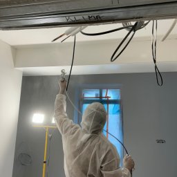 Firma ogolno budowlana Miroslaw Gros - Pracownik w kombinezonie maluje ścianę szarym kolorem za pomocą pistoletu natryskowego, widoczne kable i oświetlenie budowlane. Prace wykończeniowe w pomieszczeniu.
