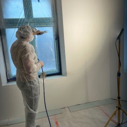 Firma ogolno budowlana Miroslaw Gros - Pracownik w kombinezonie maluje ścianę natryskowo. Okno zabezpieczone folią. Widoczny wąż od pistoletu i lampa oświetleniowa. Świeżo pomalowana ściana.