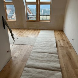 Firma ogolno budowlana Miroslaw Gros - Jasne, drewniane panele podłogowe w pokoju na poddaszu, częściowo zakryte papierem ochronnym. Widoczne okna dachowe i fragment górskiego krajobrazu.