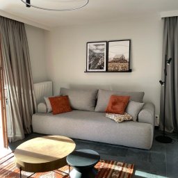 Firma ogolno budowlana Miroslaw Gros - Jasnoszara sofa z pomarańczowymi poduszkami na tle ściany z grafikami, okrągły stolik kawowy i wzorzysty dywan tworzą przytulną aranżację wnętrza.