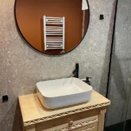 Jasna, drewniana szafka z umywalką w nowoczesnej łazience z okrągłym lustrem, na tle szarych płytek i grzejnika. Styl góralski.