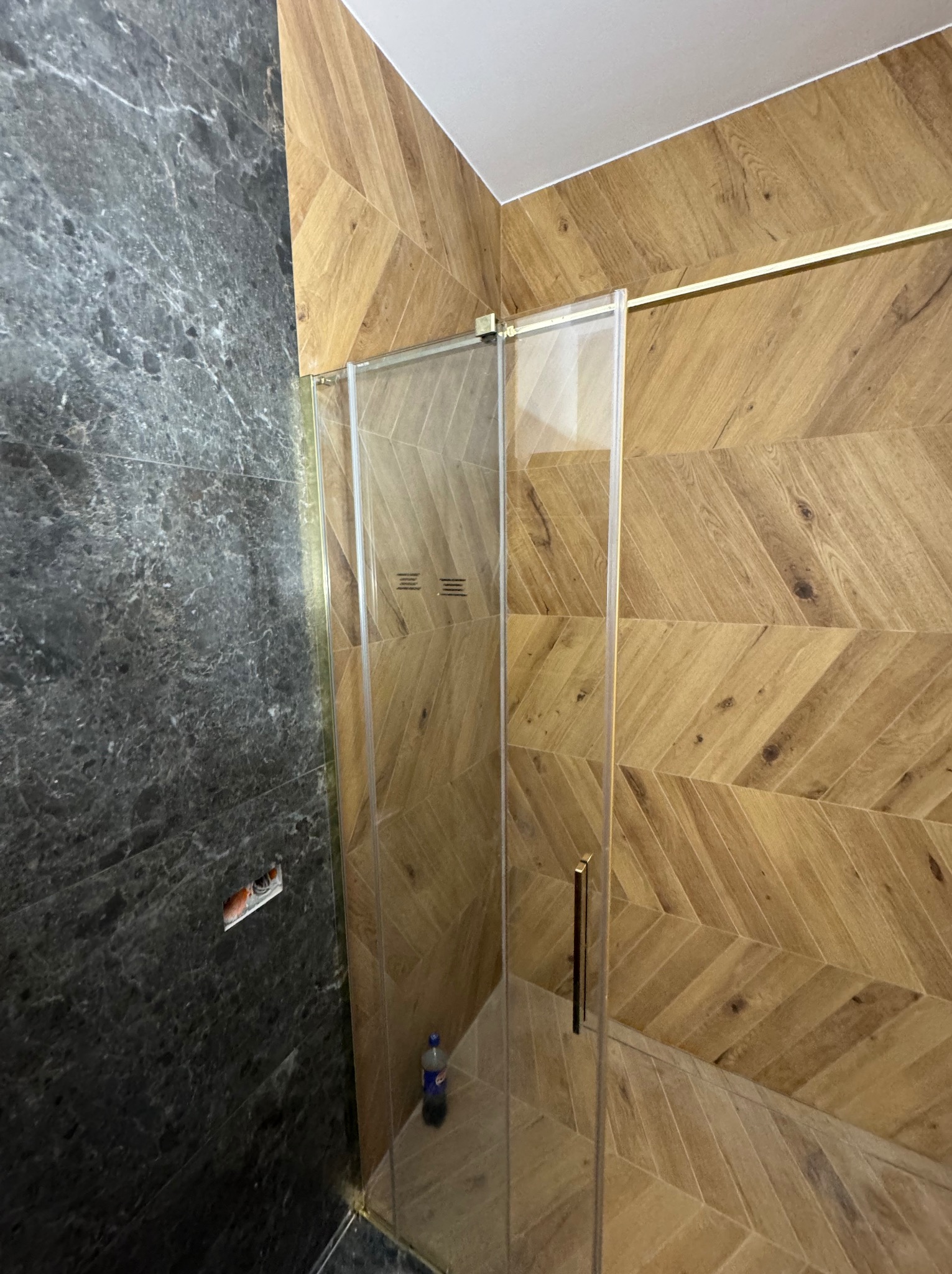 Szklana kabina prysznicowa z złotymi elementami, obłożona płytkami imitującymi drewno w jodełkę i szarym marmurem. Widoczna butelka w rogu kabiny.