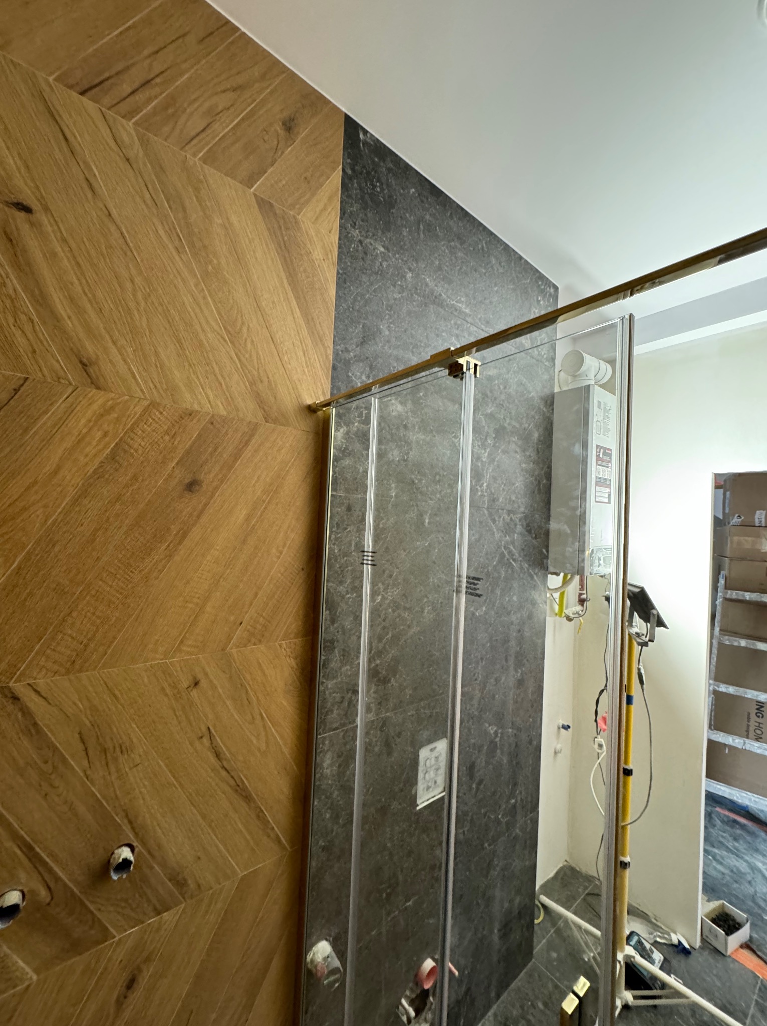 Łazienka w trakcie wykończenia: drewnopodobne płytki ułożone w jodełkę, ciemne płytki imitujące kamień, szklana kabina prysznicowa z elementami w kolorze złotym.