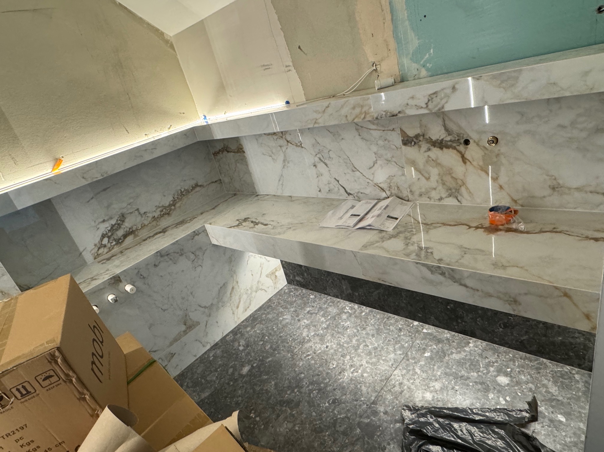 Montaż płytek imitujących marmur w łazience, widoczne fugi, oświetlenie LED, kartony i narzędzia. Prace wykończeniowe w toku, surowy stan ścian.