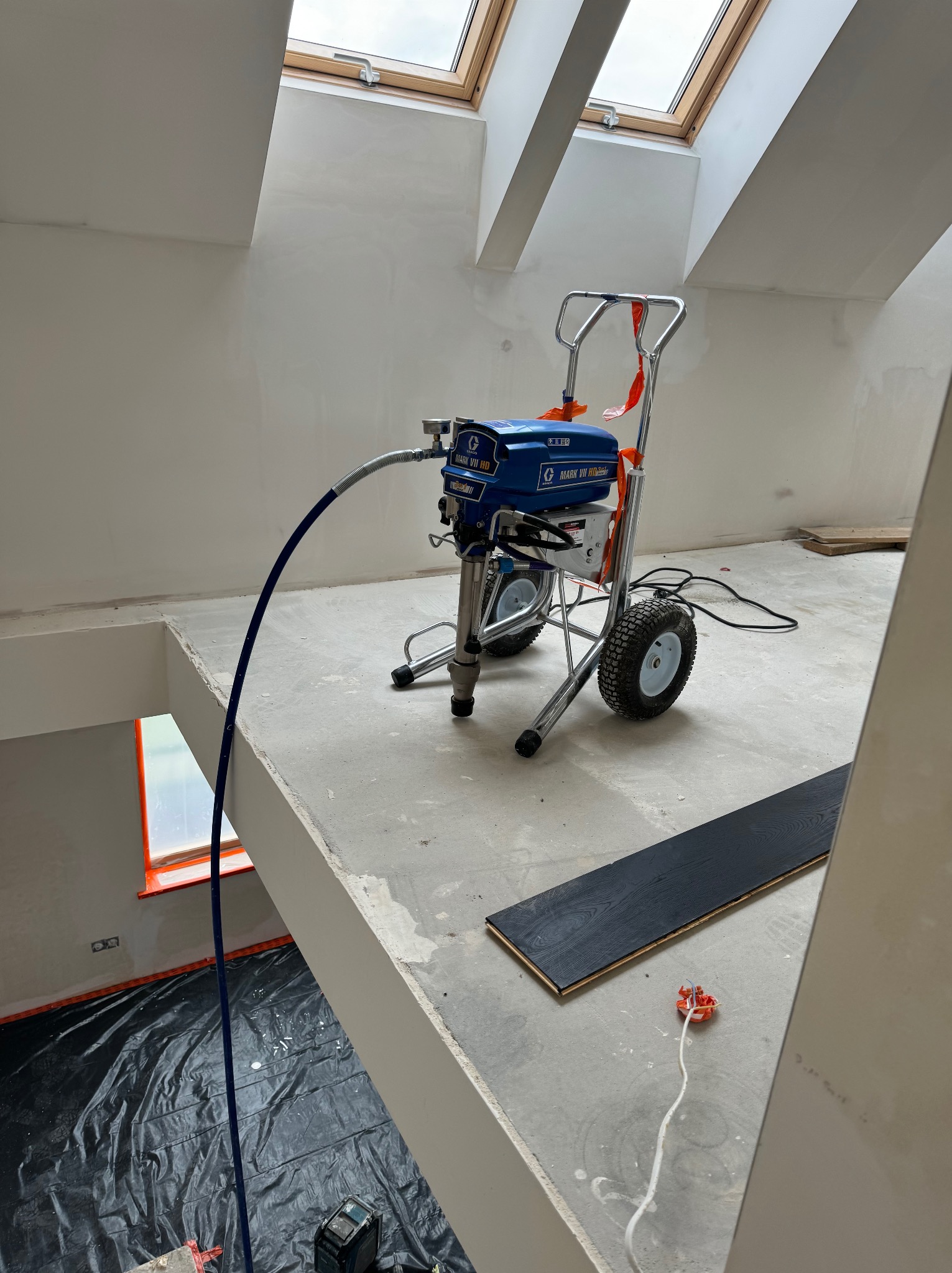 Agregat malarski na poddaszu z oknami dachowymi, przygotowany do pracy. Podłoże zabezpieczone folią. Widoczne fragmenty paneli podłogowych.