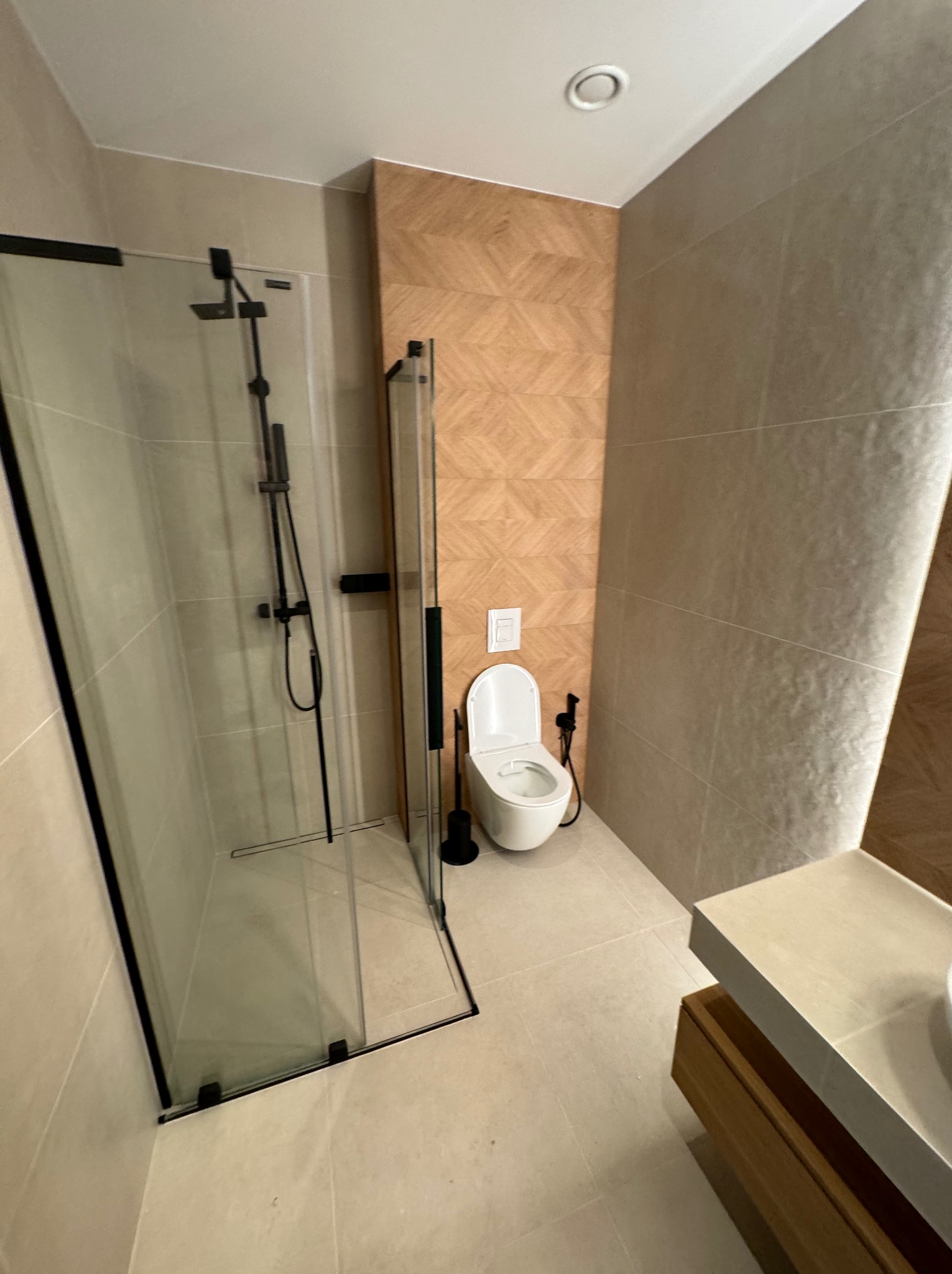 Nowoczesna łazienka z kabiną prysznicową typu walk-in z czarnymi ramami, toaletą i umywalką z drewnianą szafką. Płytki w odcieniach beżu i brązu.