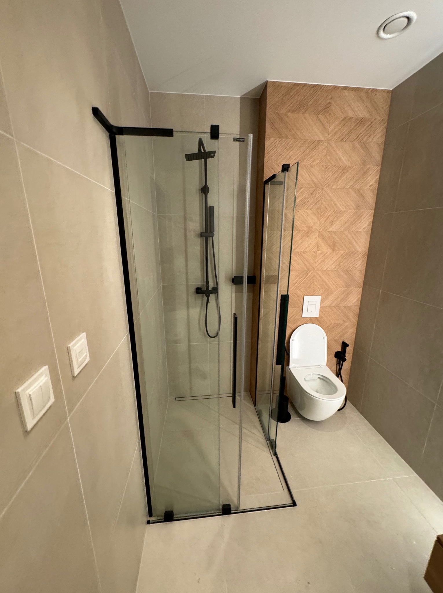 Nowoczesna łazienka z kabiną prysznicową typu walk-in z czarnymi profilami, toaletą wiszącą i geometryczną mozaiką na ścianie. Stonowana kolorystyka.