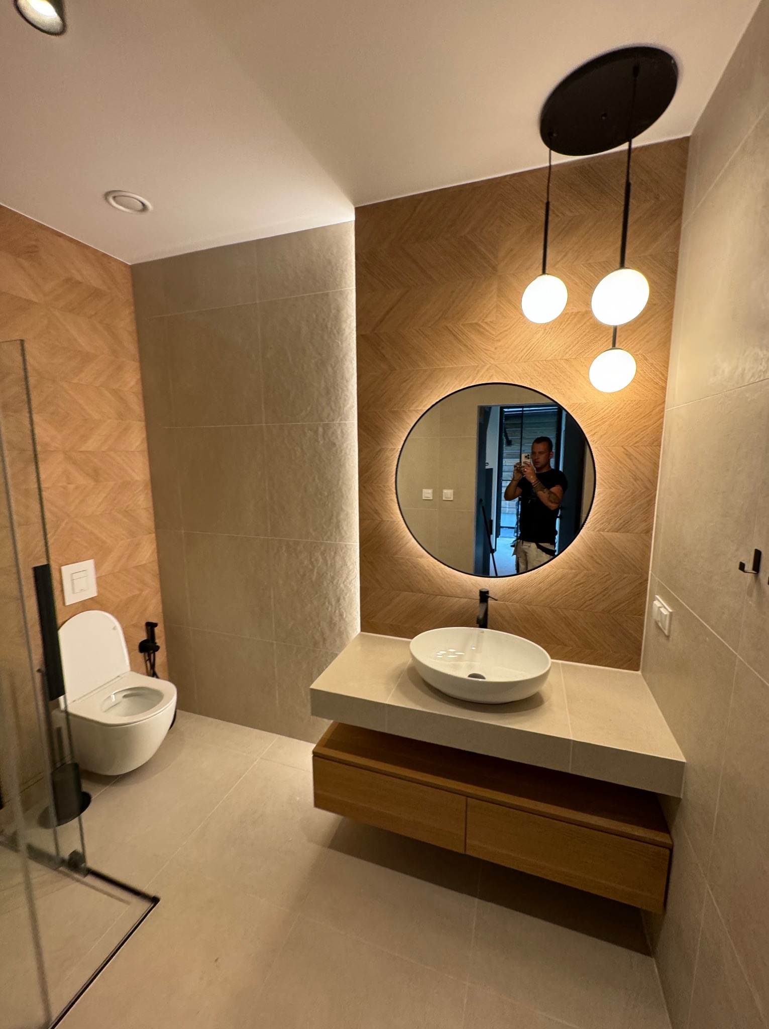 Nowoczesna łazienka z okrągłym lustrem z podświetleniem LED, wiszącą umywalką i drewnianymi szafkami. Na ścianach płytki w odcieniach beżu i brązu z wzorem jodełki.