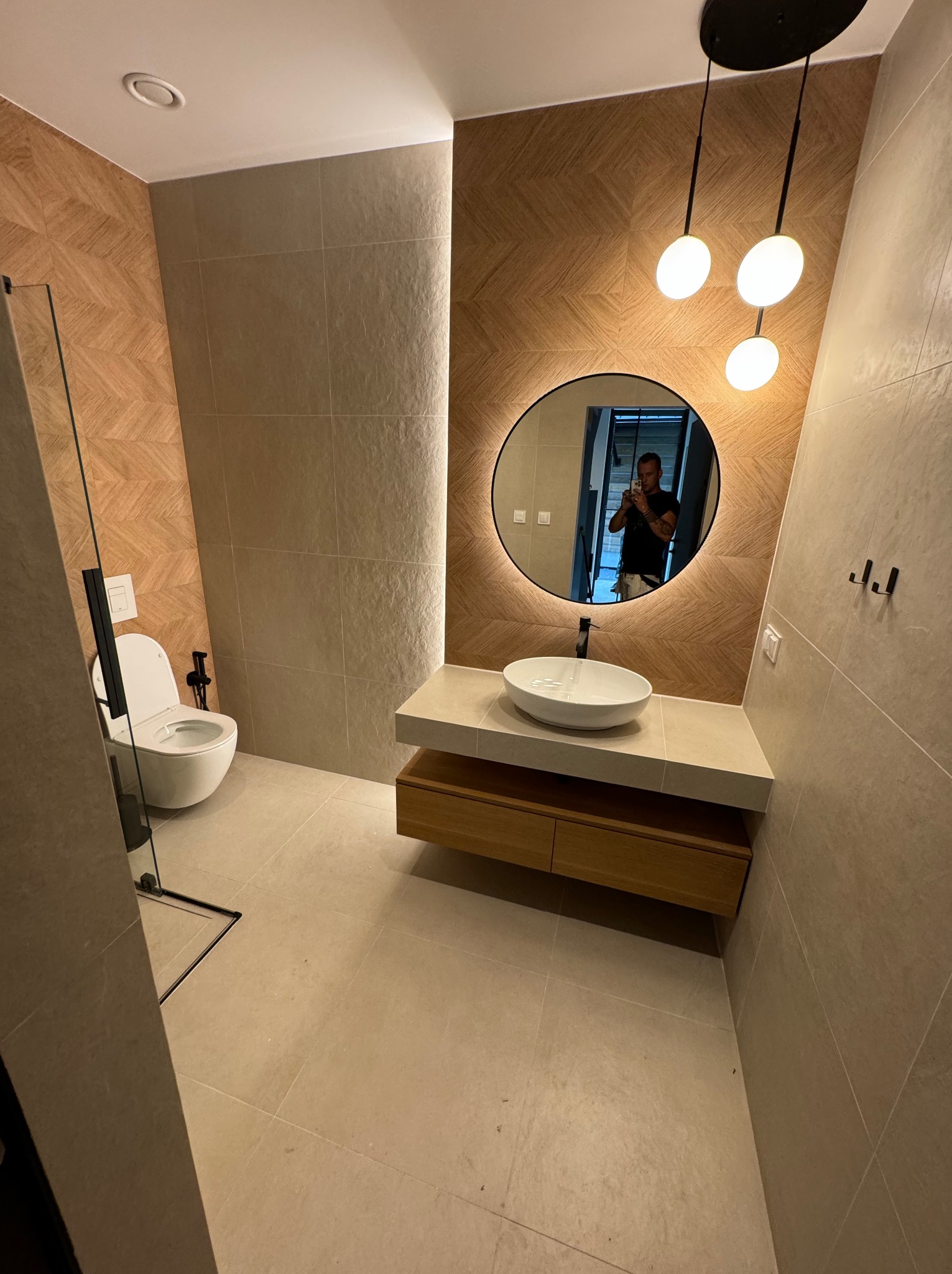 Nowoczesna łazienka z okrągłym lustrem z podświetleniem LED, drewnianą szafką pod umywalką i geometryczną mozaiką na ścianie. Widoczna kabina prysznicowa.