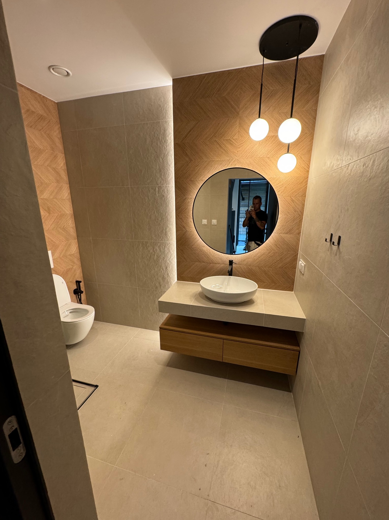 Nowoczesna łazienka z beżowymi płytkami, okrągłym lustrem z podświetleniem LED, wiszącą szafką z umywalką i podwieszaną toaletą. Na ścianie wieszaki.
