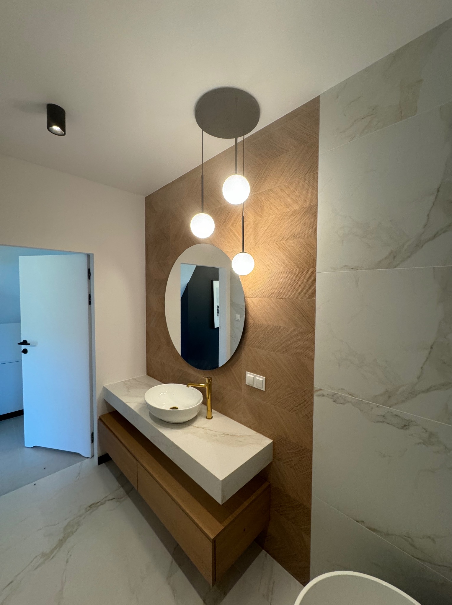 Nowoczesna łazienka z okrągłym lustrem, drewnianą szafką pod umywalką i marmurową posadzką. Dekoracyjna ściana z drewnopodobnych paneli.