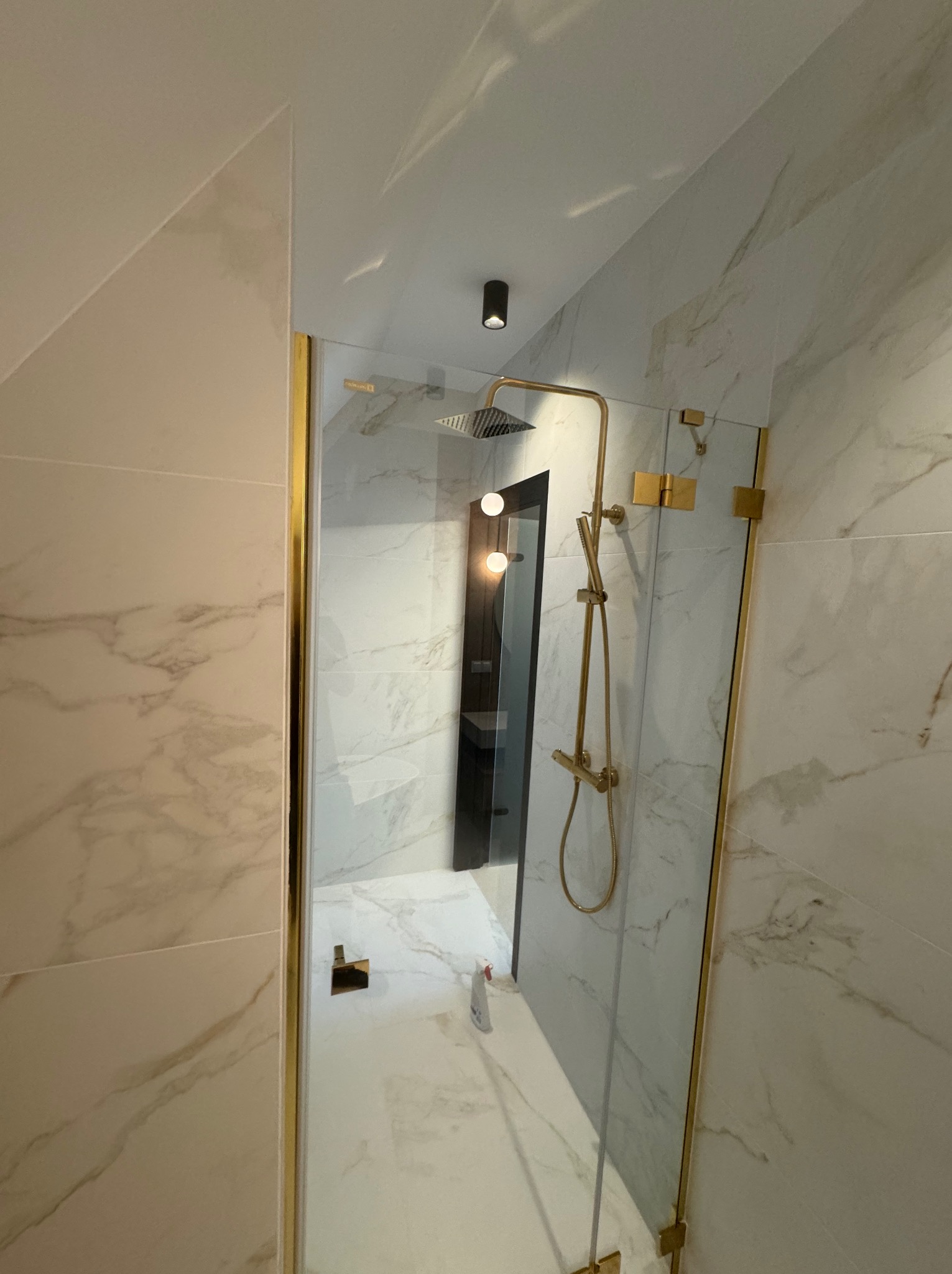 Elegancka kabina prysznicowa z marmurowymi płytkami i złotymi elementami wykończeniowymi. Widoczny deszczownica i bateria w kolorze złota.