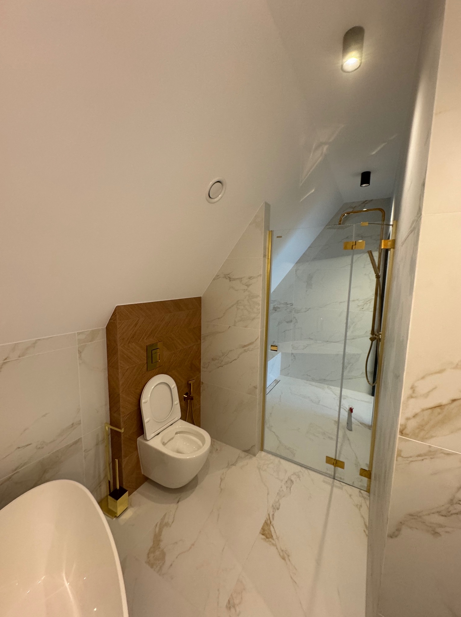 Nowoczesna łazienka na poddaszu z marmurowymi płytkami, złotymi akcentami i kabiną prysznicową typu walk-in. Uwagę zwraca nietypowa zabudowa toalety.