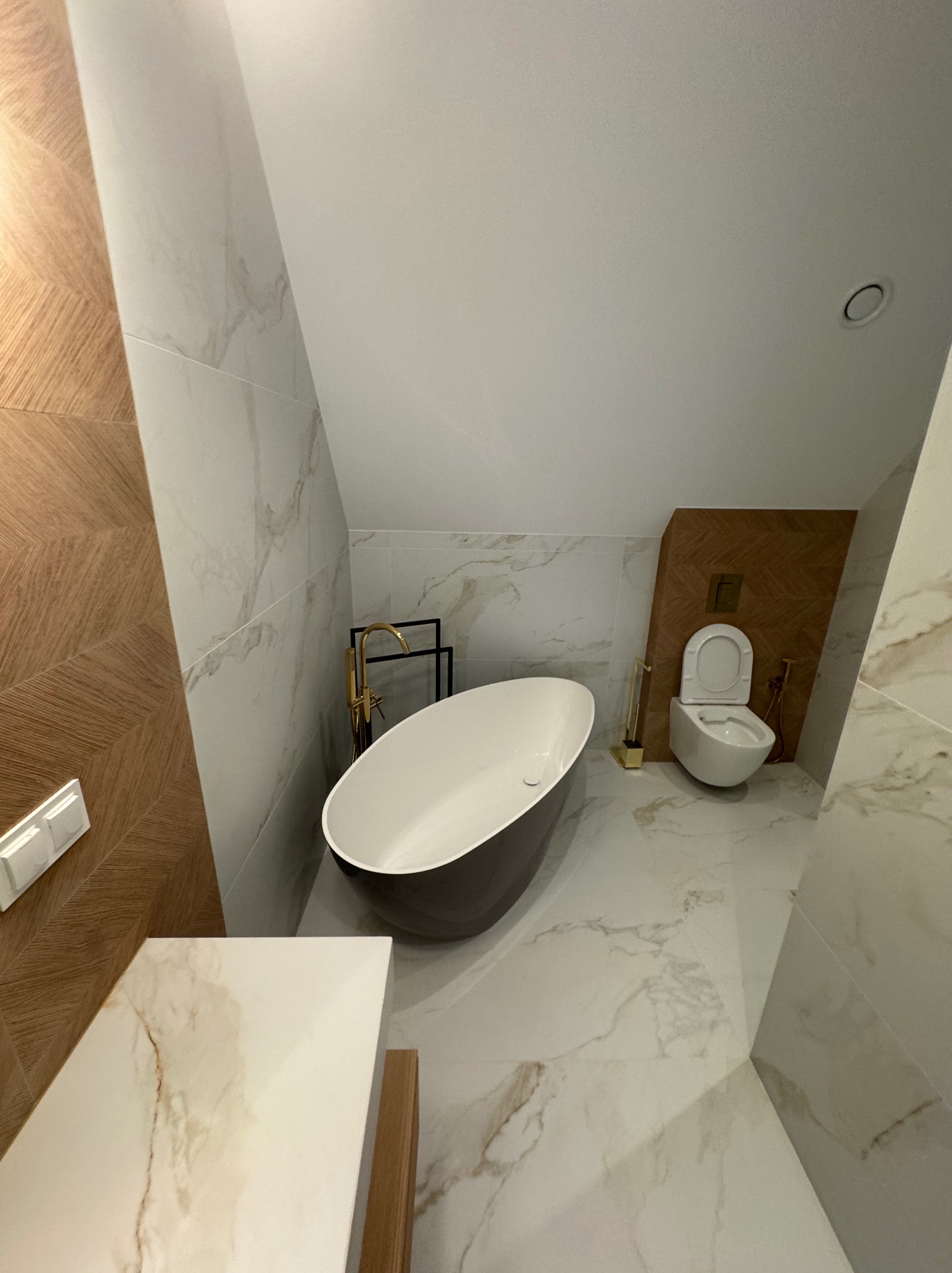 Elegancka łazienka na poddaszu z wolnostojącą wanną, złotą armaturą i płytkami imitującymi marmur. Uwagę zwraca drewniana ściana z wzorem jodełki.