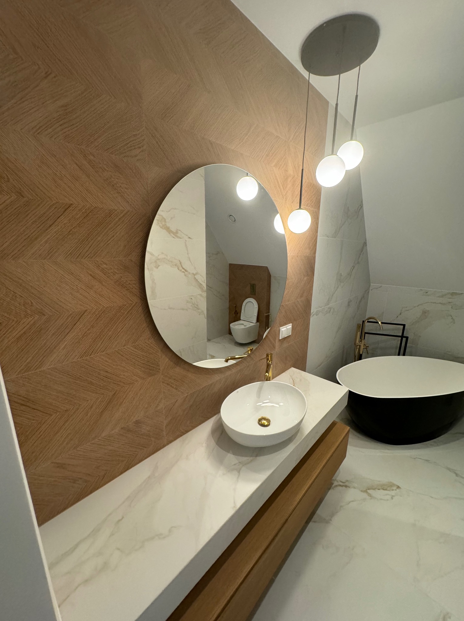 Nowoczesna łazienka z owalnym lustrem, umywalką na blacie z marmuru i wolnostojącą wanną. Na ścianie płytki w jodełkę, a w lustrze odbicie toalety.