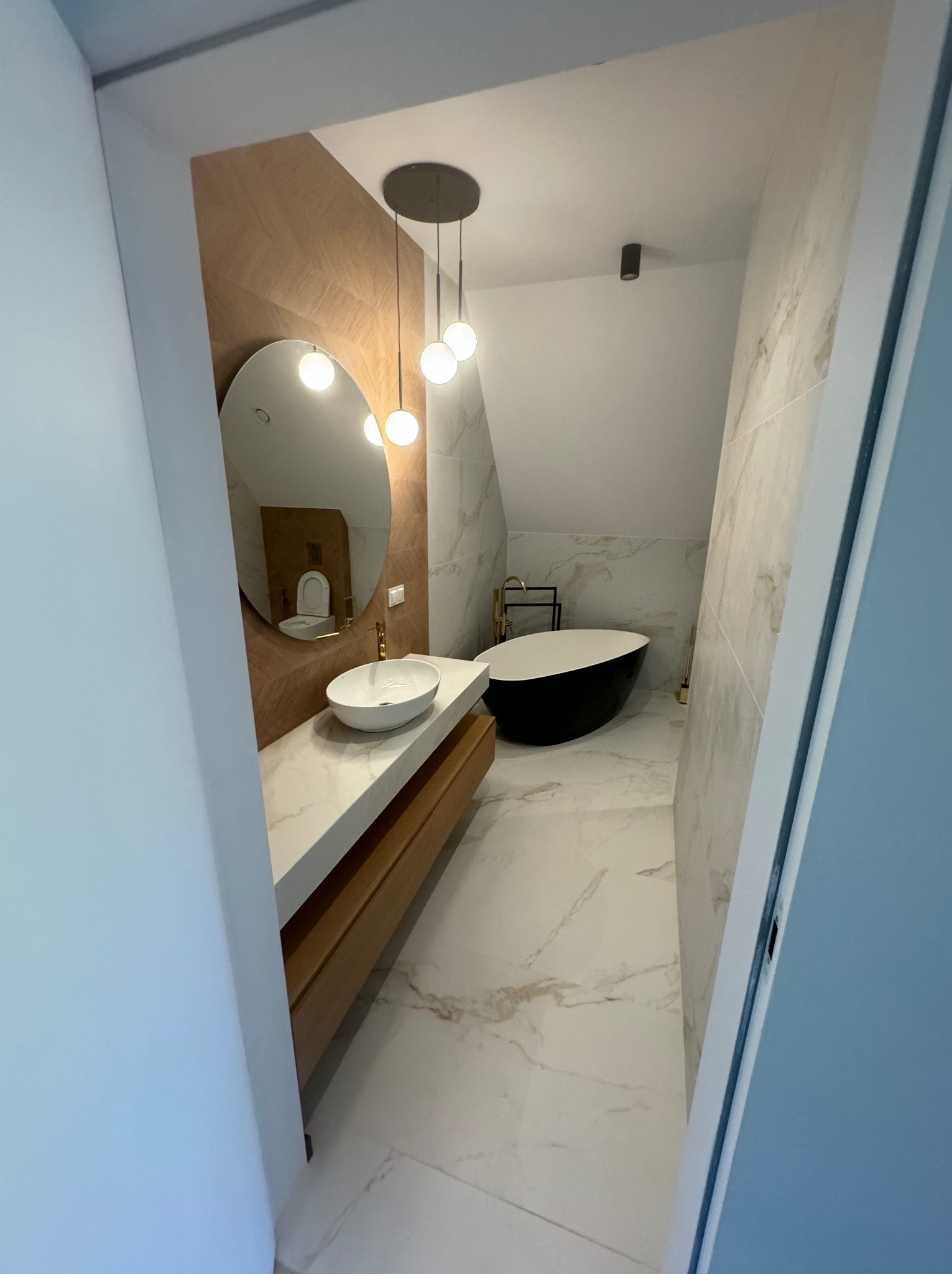 Elegancka łazienka na poddaszu z owalną wanną, marmurową posadzką i umywalką na blacie. W tle lustro odbija toaletę, a nad umywalką zwisają dekoracyjne, okrągłe lampy.