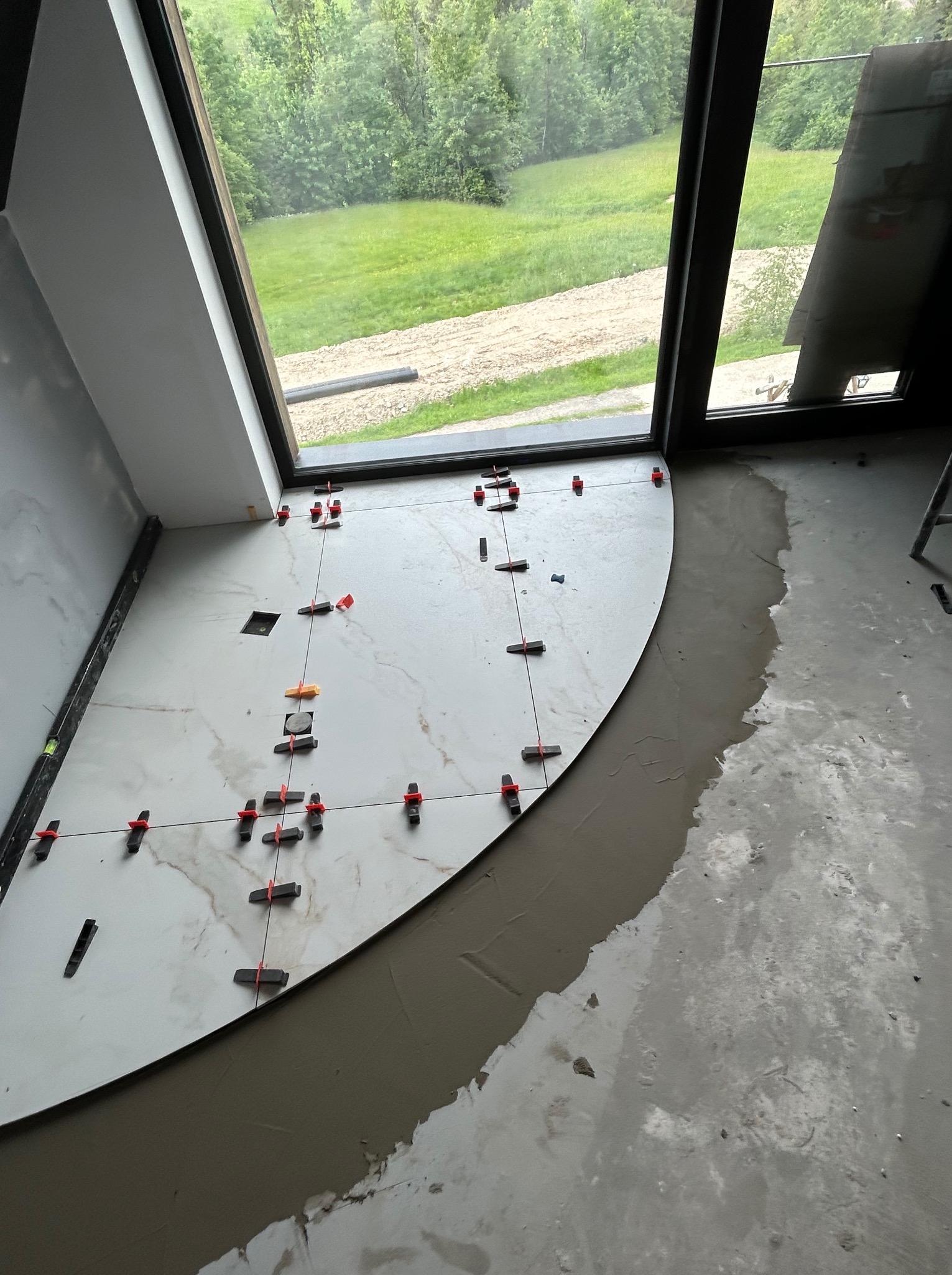 Układanie płytek wielkoformatowych z użyciem klipsów poziomujących na podłodze przy oknie z widokiem na zieleń. Częściowo nałożony klej do płytek.