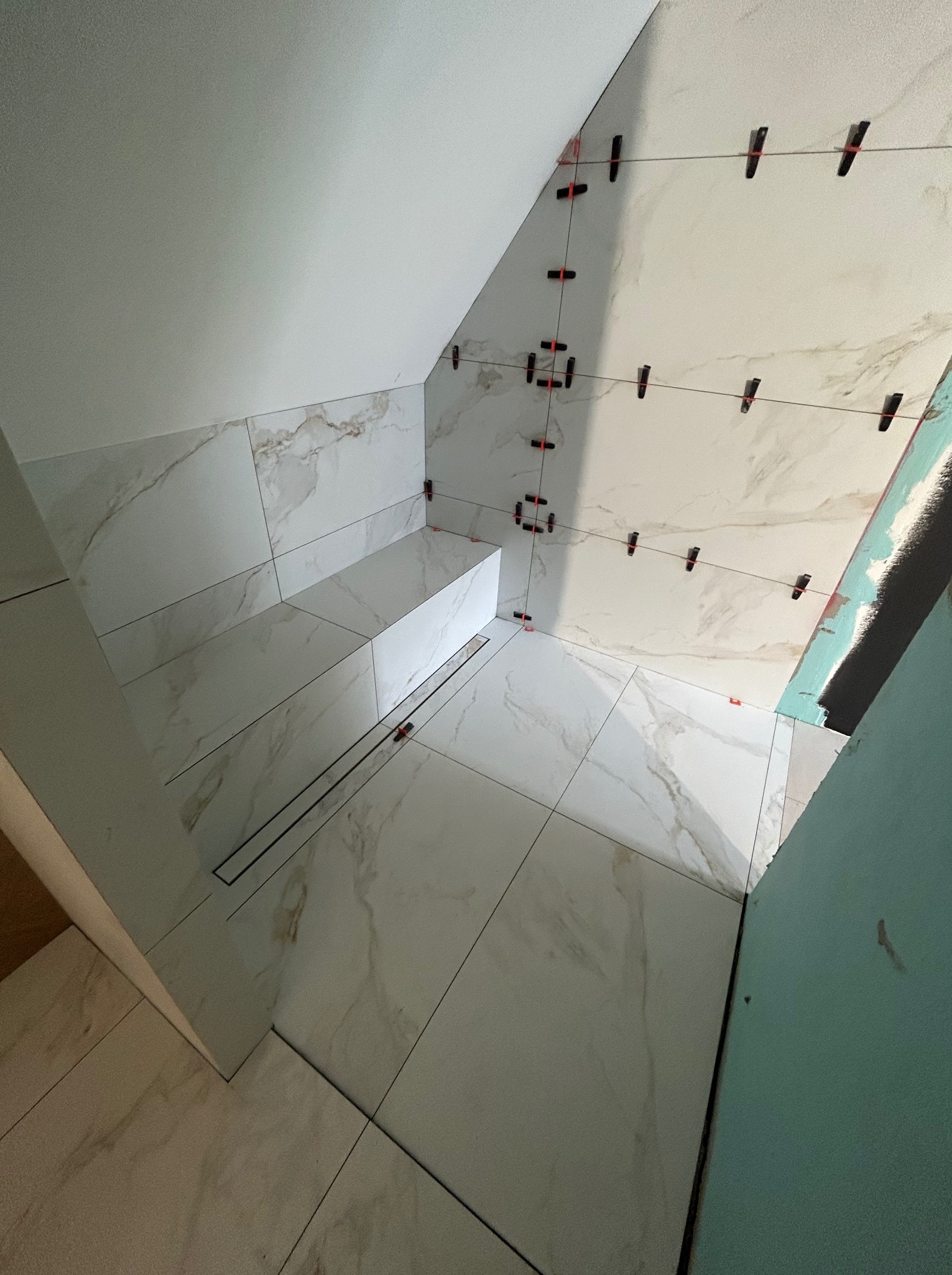 Łazienka w trakcie wykańczania: białe płytki imitujące marmur na ścianach i podłodze, system poziomowania płytek, widoczny odpływ liniowy, skos dachu.