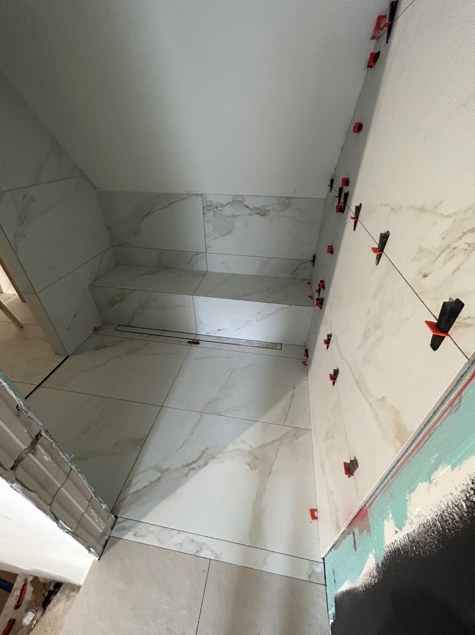 Wykończenie łazienki: białe płytki imitujące marmur na ścianach i podłodze, z systemem poziomowania płytek. Widoczny odpływ liniowy i fragment przygotowanej ściany.