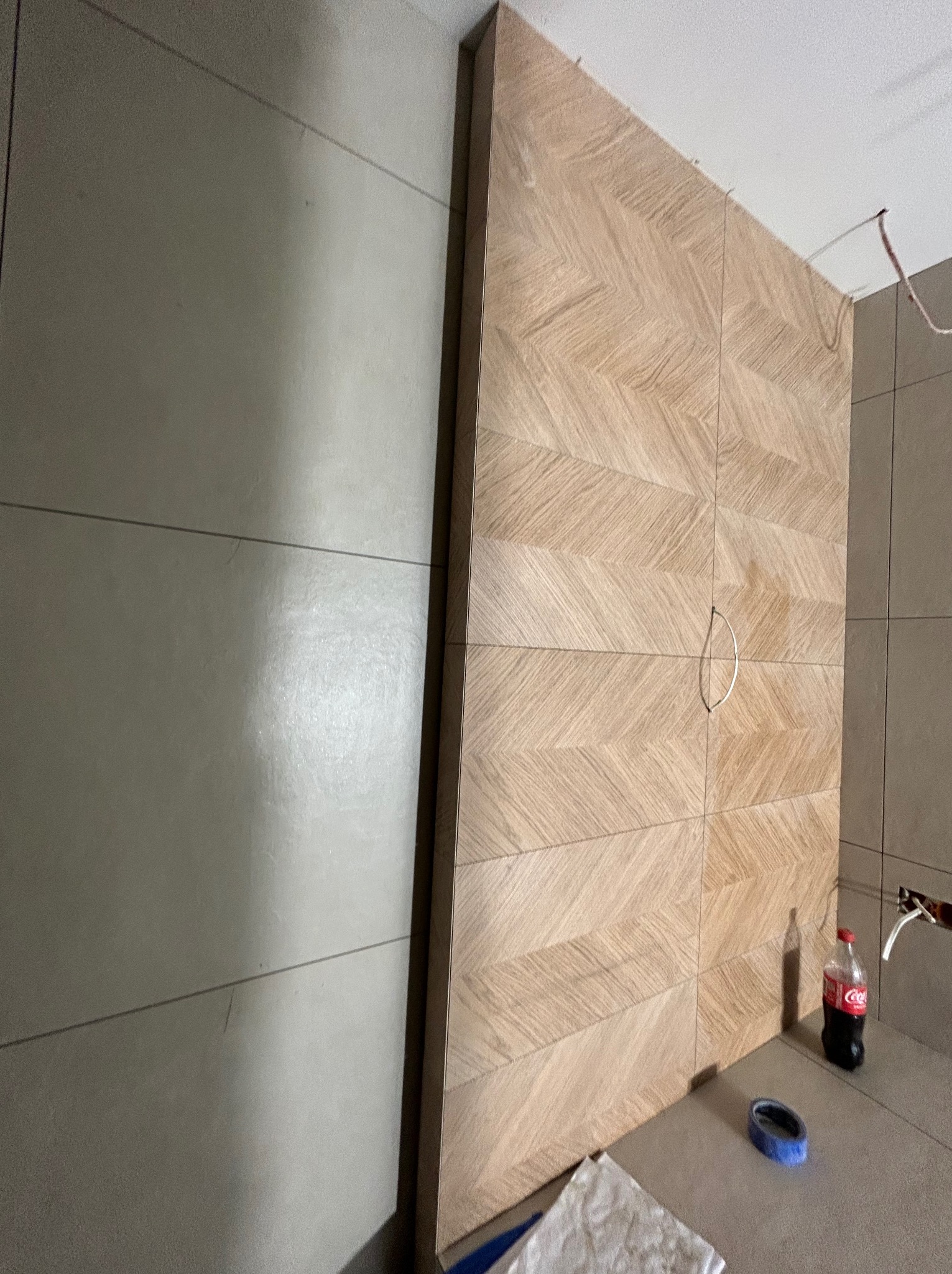 Wykończona ściana z płytek imitujących drewno ułożonych w jodełkę, z metalowym uchwytem. Na podłodze butelka napoju i taśma malarska. Widoczne płytki na ścianie obok.