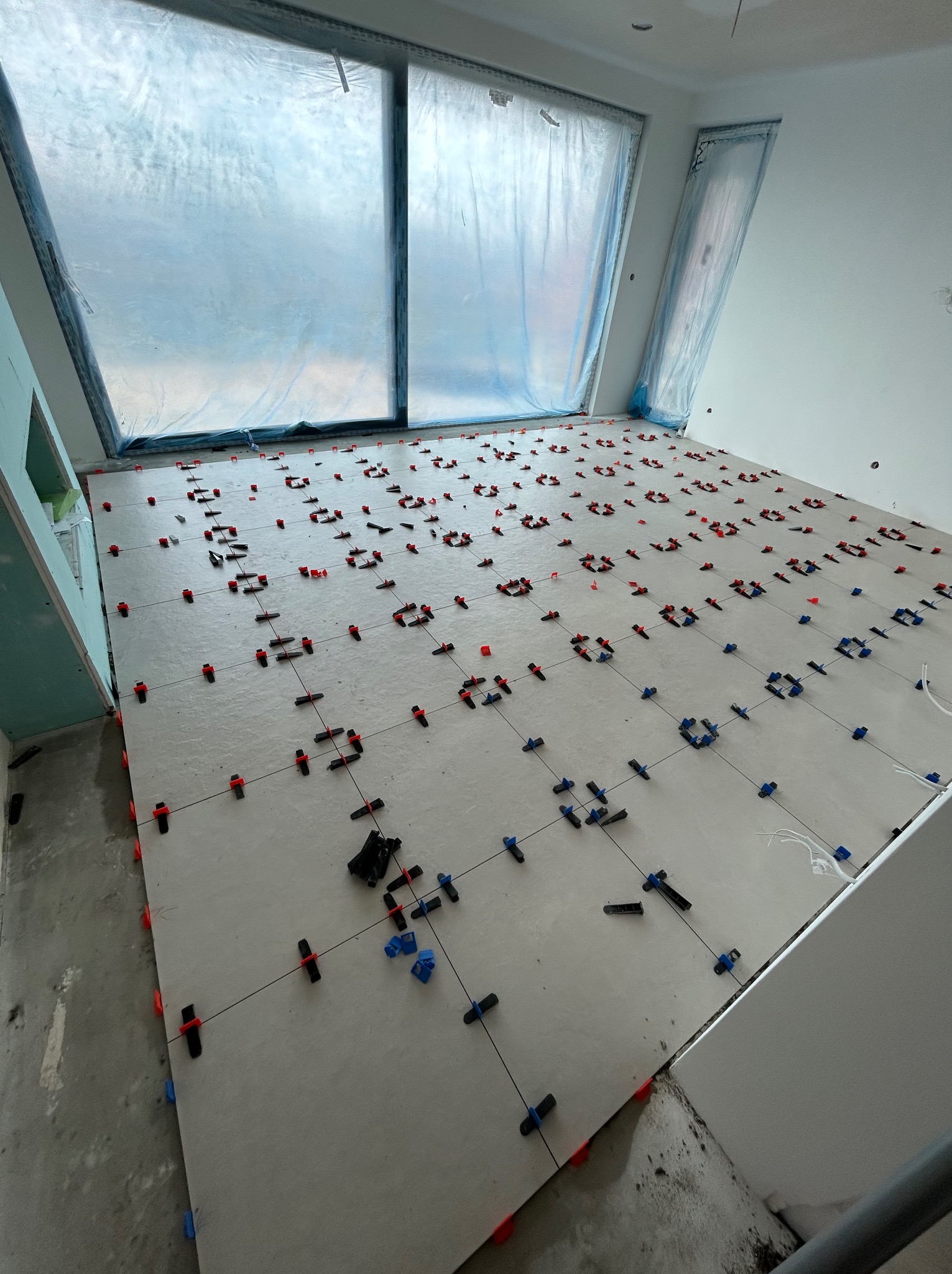 Układanie jasnych płytek podłogowych w pomieszczeniu z oknami zabezpieczonymi folią. Użyto klipsów poziomujących w kolorach czerwonym i niebieskim.