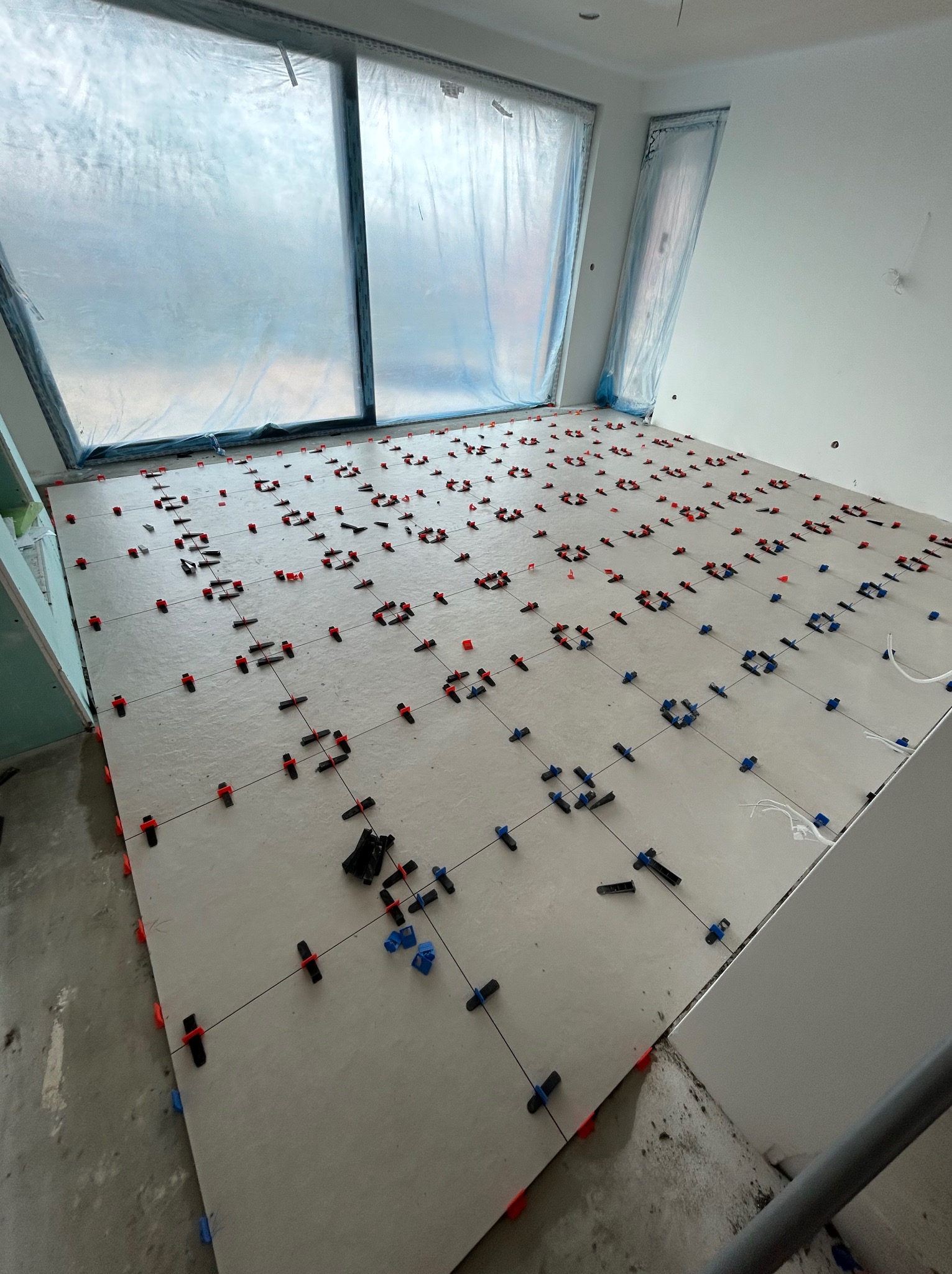 Układanie płytek podłogowych w pokoju z oknami zabezpieczonymi folią. Widoczne krzyżyki dystansowe i system poziomowania płytek. Jasne płytki, szare fugi.