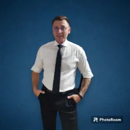 Mężczyzna w okularach, białej koszuli i czarnym krawacie, z tatuażem na lewym przedramieniu, pozuje na tle teksturowanej, niebieskiej ściany.