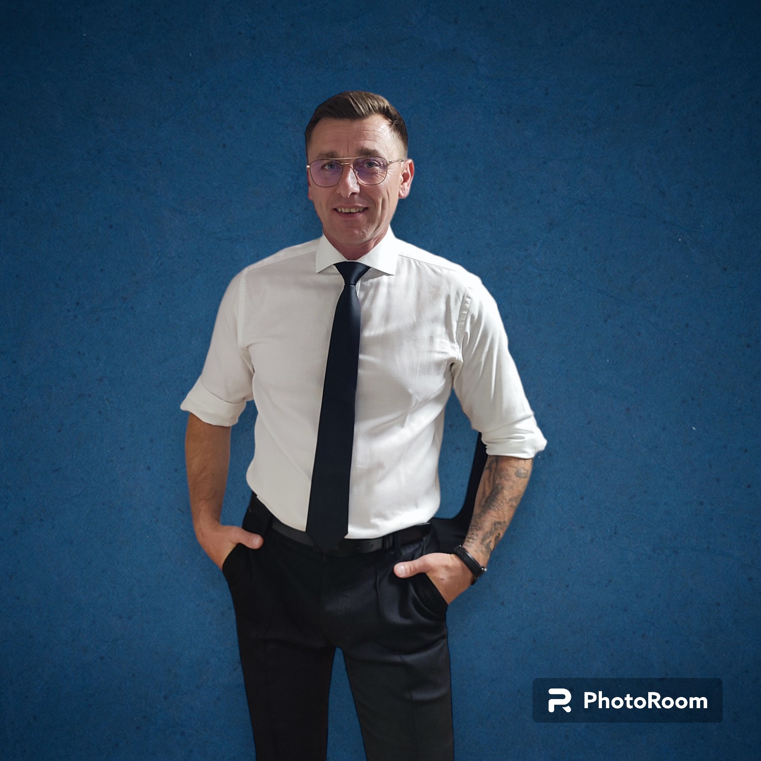 Mężczyzna w okularach, białej koszuli i czarnym krawacie, z tatuażem na lewym przedramieniu, pozuje na tle teksturowanej, niebieskiej ściany.