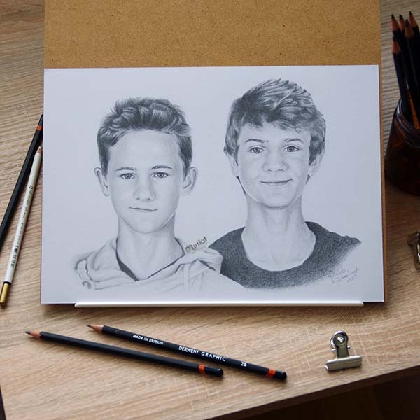Portret ołówkiem dwóch młodych mężczyzn, wykonany na białym papierze, z widocznymi ołówkami Derwent Graphic i spinaczem biurowym na drewnianym stole.