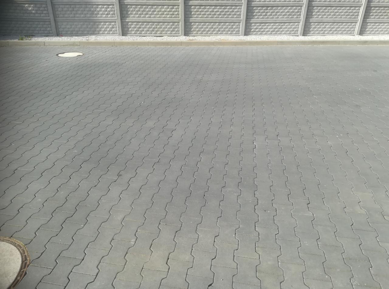 Szara kostka brukowa ułożona w charakterystyczny wzór na dużej powierzchni, z betonowym ogrodzeniem panelowym w tle i włazem kanalizacyjnym w rogu kadru.