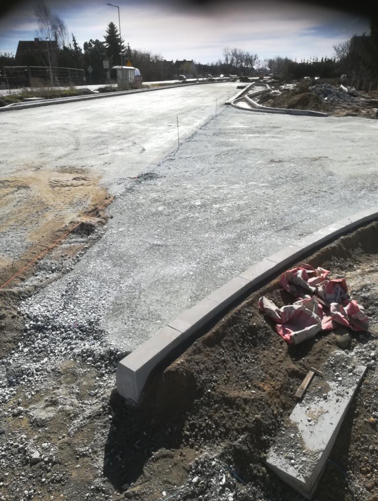 Przygotowanie podłoża pod nawierzchnię drogową z ułożonymi obrzeżami, widoczne warstwy kruszywa i piasku, narzędzia oraz worki na ziemi wzdłuż krawężnika.