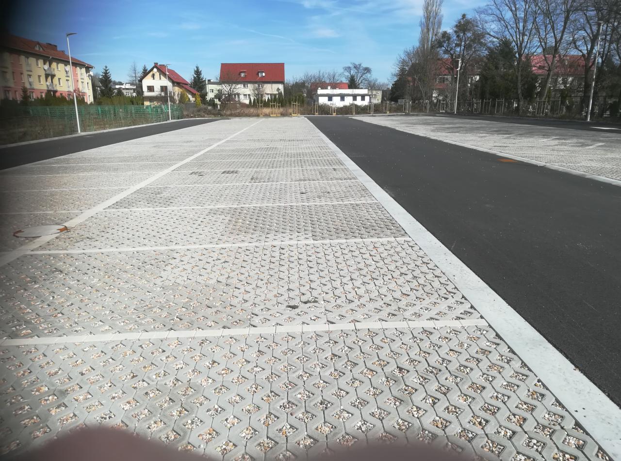 Nowo wybudowany parking z ażurowych płyt betonowych wypełnionych drobnym kruszywem, oddzielony pasem asfaltu, w tle budynki mieszkalne pod błękitnym niebem.