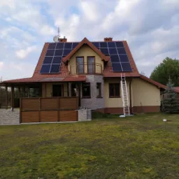 Dom jednorodzinny z panelami fotowoltaicznymi na dachu z czerwonej dachówki, drabina oparta o ścianę, widok z ogrodu w pochmurny dzień.