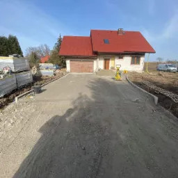 Przygotowanie podjazdu do domu z czerwonym dachem: ubijanie podłoża wibratorem, obrzeża ułożone, palety z kostką czekają na montaż.