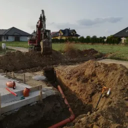 Czerwona koparka wykonuje wykop pod instalację rur kanalizacyjnych na działce budowlanej, widoczny człowiek w tle oraz narzędzia i materiały budowlane.