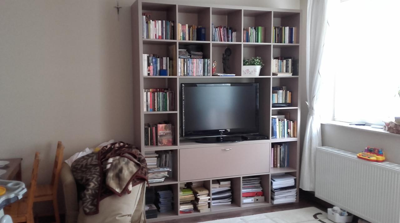 Duża, beżowa szafa na wymiar z półkami na książki i wnęką na telewizor, w tle okno z białą zasłoną i grzejnik.