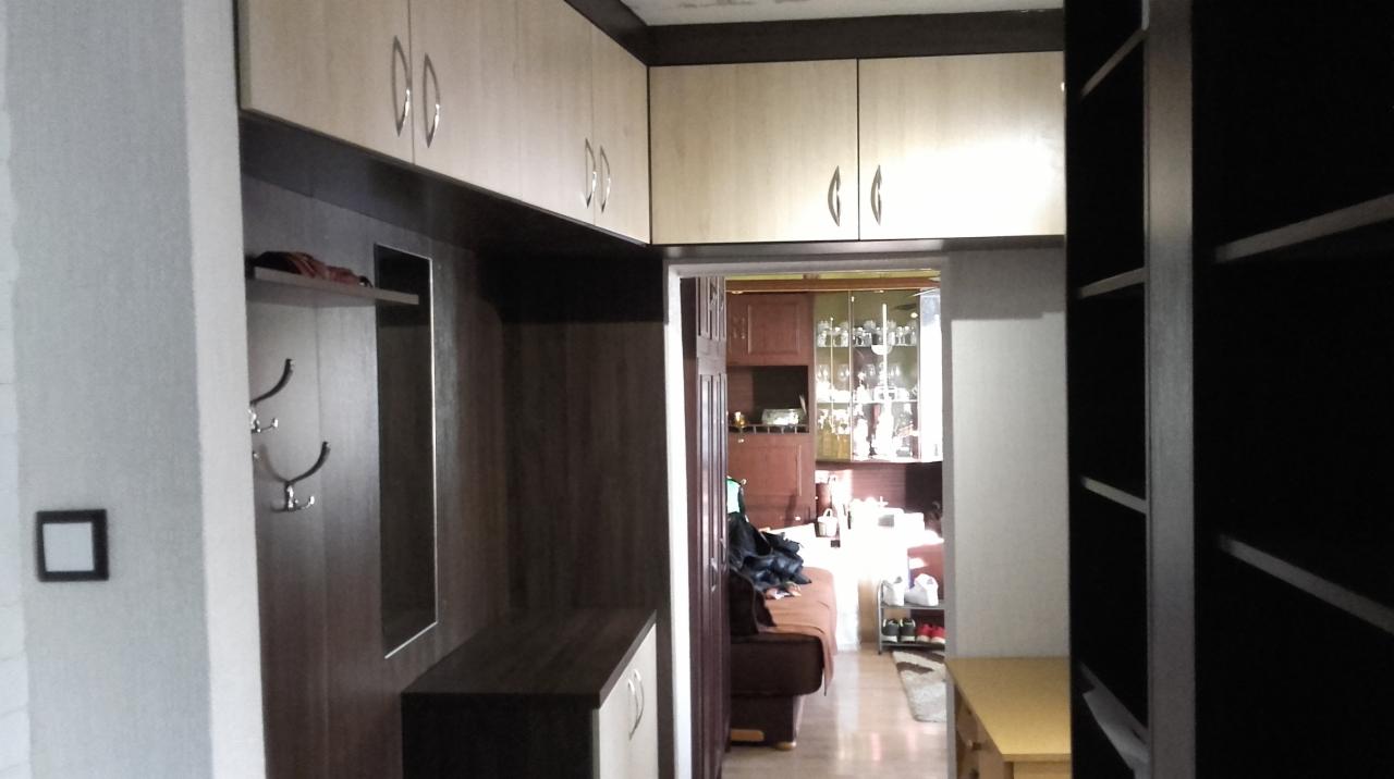 Przedpokój z zabudową meblową: szafki górne w kolorze jasnego drewna, ciemnobrązowy panel z wieszakami i lustrem, widok na wnętrze mieszkania z meblami i dekoracjami.