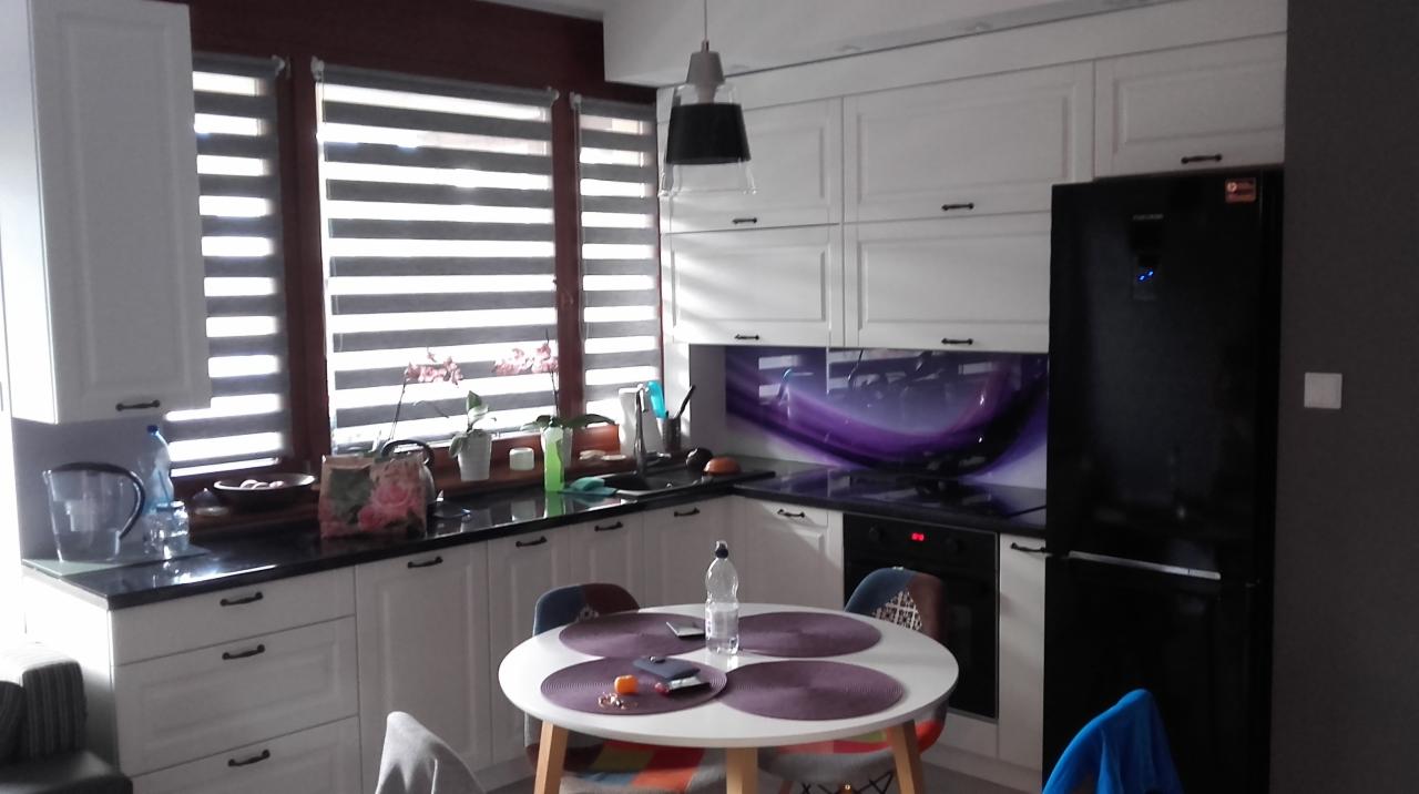 Jasna kuchnia z białymi szafkami, czarnym blatem i kolorowym panelem nad kuchenką, okrągły stół z czterema krzesłami na pierwszym planie.