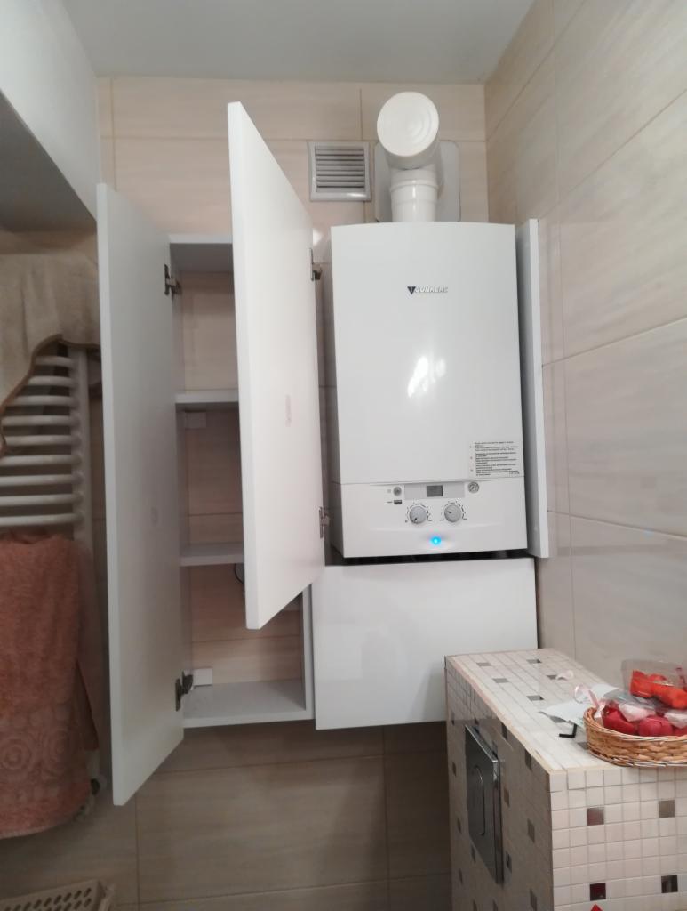 Biały piec gazowy marki Junkers zabudowany w szafce łazienkowej z otwartymi drzwiczkami, widoczna kratka wentylacyjna i fragment grzejnika z ręcznikiem.