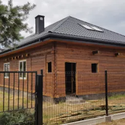 Drewniany dom w trakcie budowy z czarnym dachem z dachówki, widoczne otwory okienne i drzwiowe, ogrodzenie z siatki, drzewo iglaste po lewej stronie.