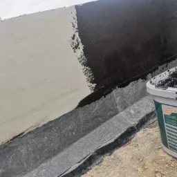 Hydroizolacja fundamentu: aplikacja czarnej, bitumicznej masy uszczelniającej na ścianę fundamentową, widoczny fragment ściany z nałożoną warstwą oraz wiadro z produktem PCI Pecimor 2K.