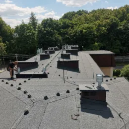 Pracownik w czarnej koszulce montuje element na płaskim dachu pokrytym papą, widoczne kominki wentylacyjne, zabezpieczenia linowe i narzędzia