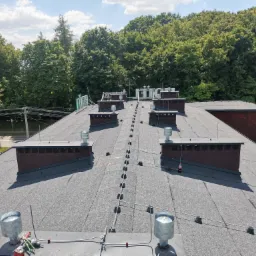 Widok z góry na płaski dach pokryty papą z licznymi kominkami wentylacyjnymi i zabezpieczeniami przeciwśniegowymi, na tle zielonych drzew i błękitnego nieba.