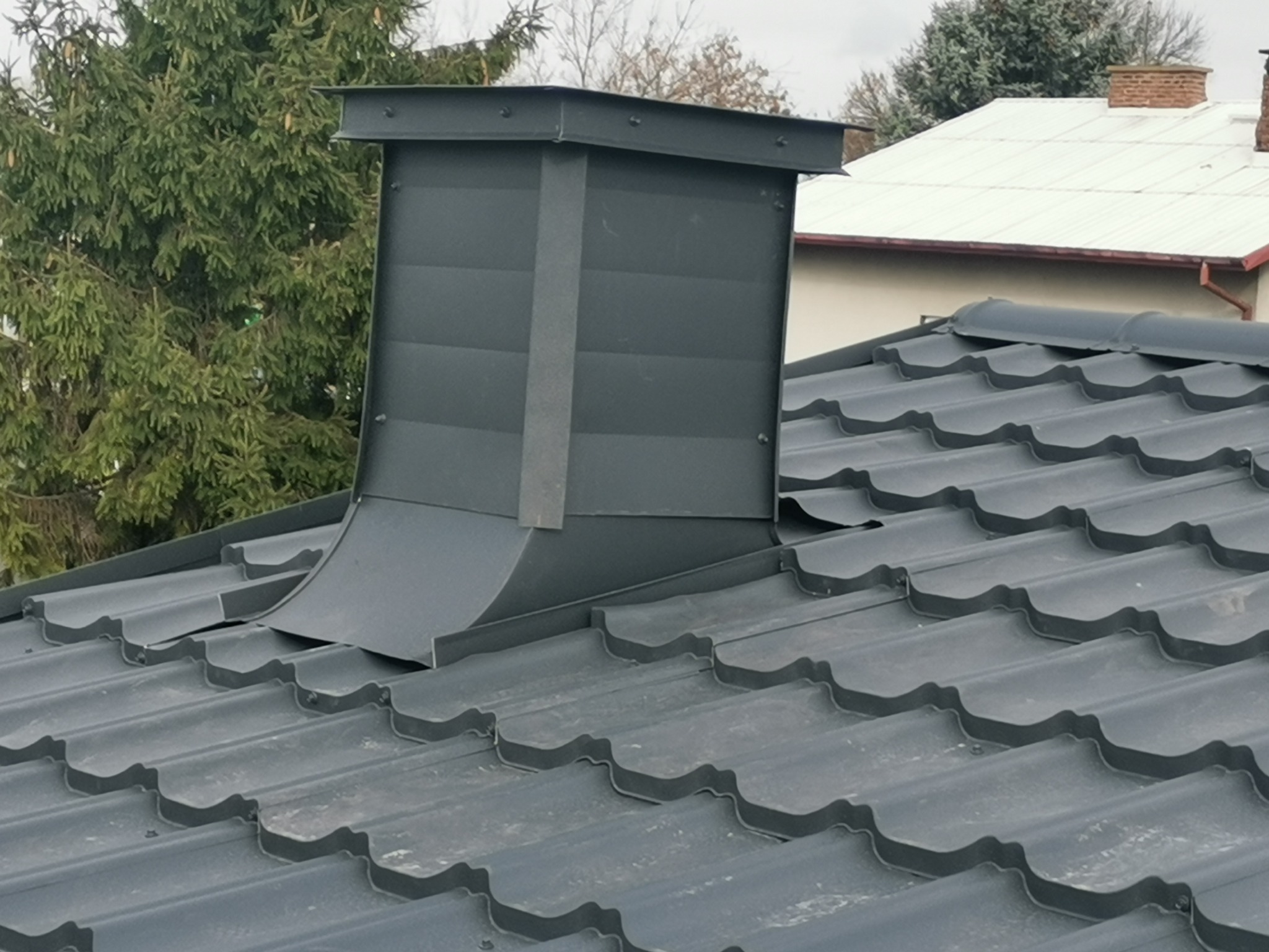 Nowoczesny, grafitowy komin wentylacyjny z blachy na dachu krytym szarą blachodachówką, widoczne połączenia blach nitami.