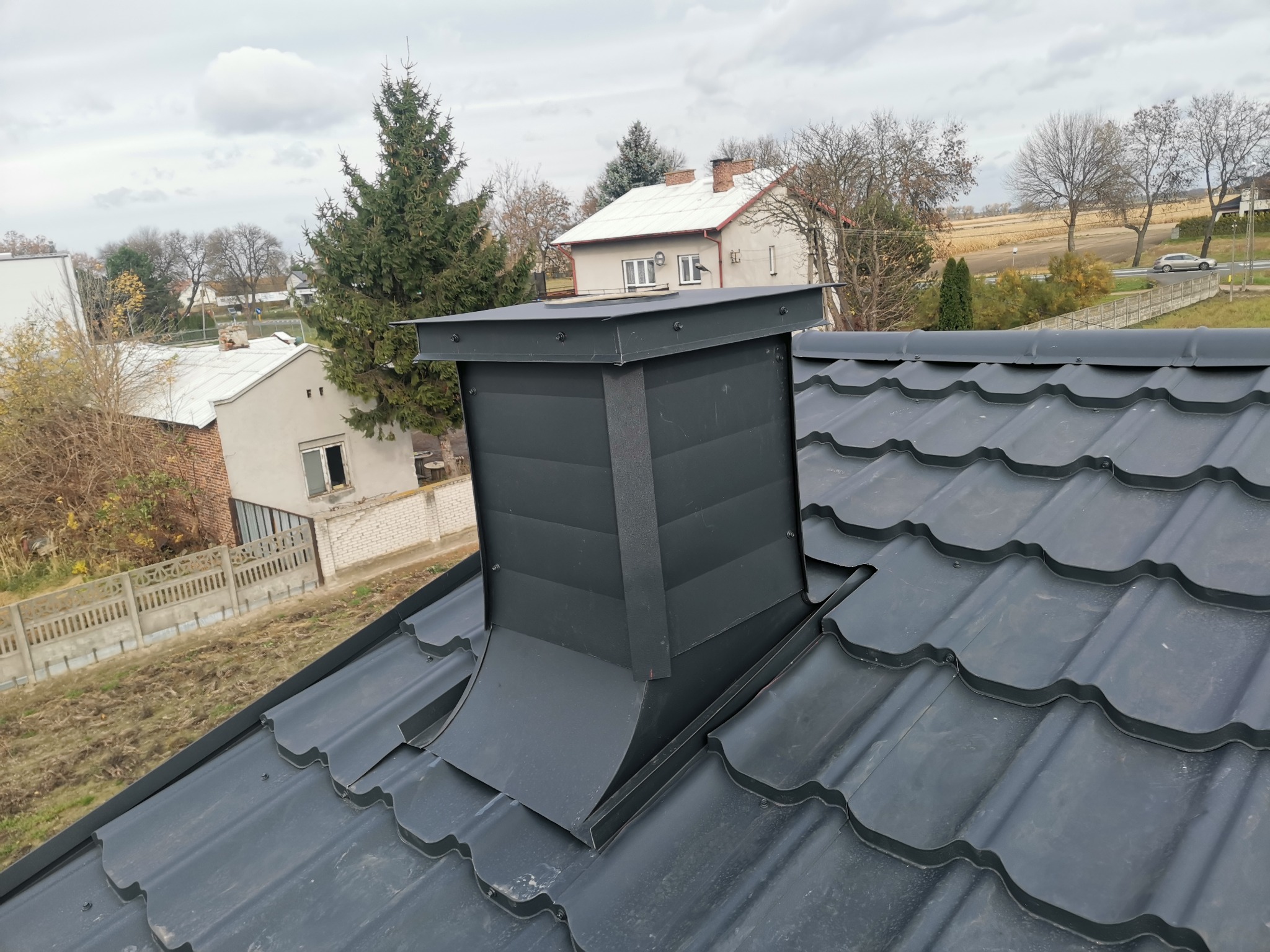 Nowy, czarny komin wentylacyjny z blachy na dachu z ciemnoszarej blachodachówki modułowej, w tle zabudowa jednorodzinna i pochmurne niebo.