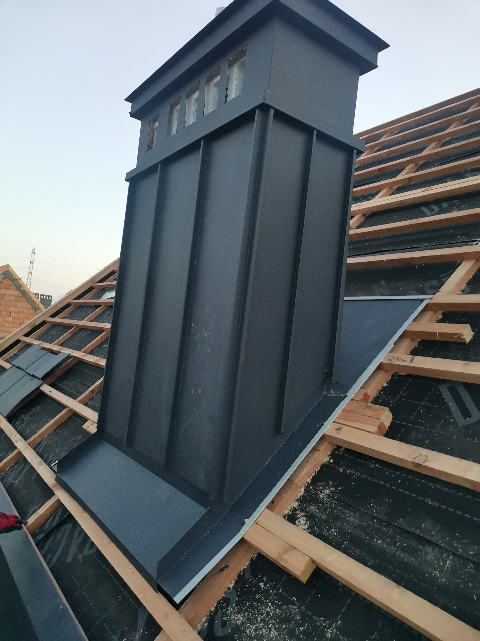 Czarny, metalowy komin z okienkami na dachu w trakcie budowy, widoczne drewniane krokwie i membrana dachowa, w tle ceglany budynek.