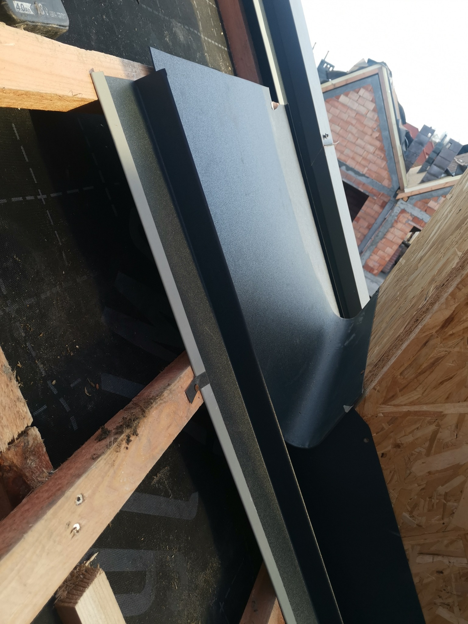 Montaż obróbki blacharskiej na dachu z widoczną membraną dachową i konstrukcją więźby dachowej, w tle fragment budynku w trakcie budowy.