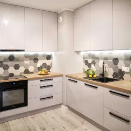 Biała, nowoczesna kuchnia z drewnianym blatem, heksagonalnymi płytkami na ścianie i czarną armaturą, oświetlenie podszafkowe LED, widok na piekarnik i zlew.